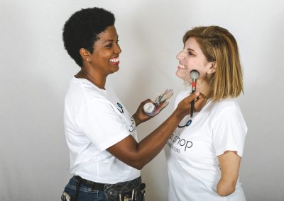 Imagen de una mujer joven de raza negra que mira de frente mientras la maquilla a otra de raza blanca que tiene amputado un brazo por debajo del codo. Ambas están sonriendo y lucen una camiseta blanca con el logotipo de Dadai Shop en el pecho.