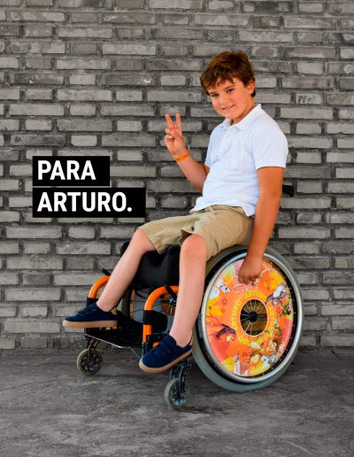 Imagen de un niño luciendo zapatillas Timper. Sonríe, va en silla de ruedas y hace el gesto de victoria con los dedos. La foto está rotulada con la frase: Para Arturo.
